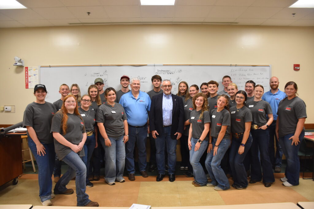 A large group of students and instructors, mostly wearing matching grey t-shirts or blue polo shirts, pose for a group photo in front of a whiteboard in a classroom.