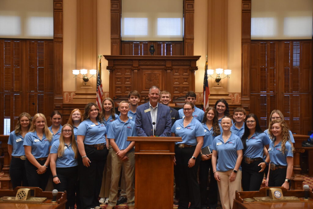 A group of students in matching light blue polo shirts pose with a man standing at a wooden podium inside a government chamber with ornate wooden paneling and flags.