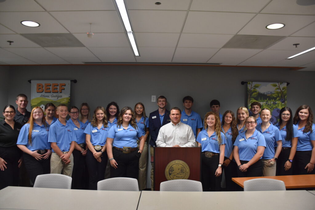A large group of students in light blue polo shirts pose indoors around Georgia Agriculture Commissioner Tyler Harper, who is standing at a podium bearing the Georgia Department of Agriculture seal