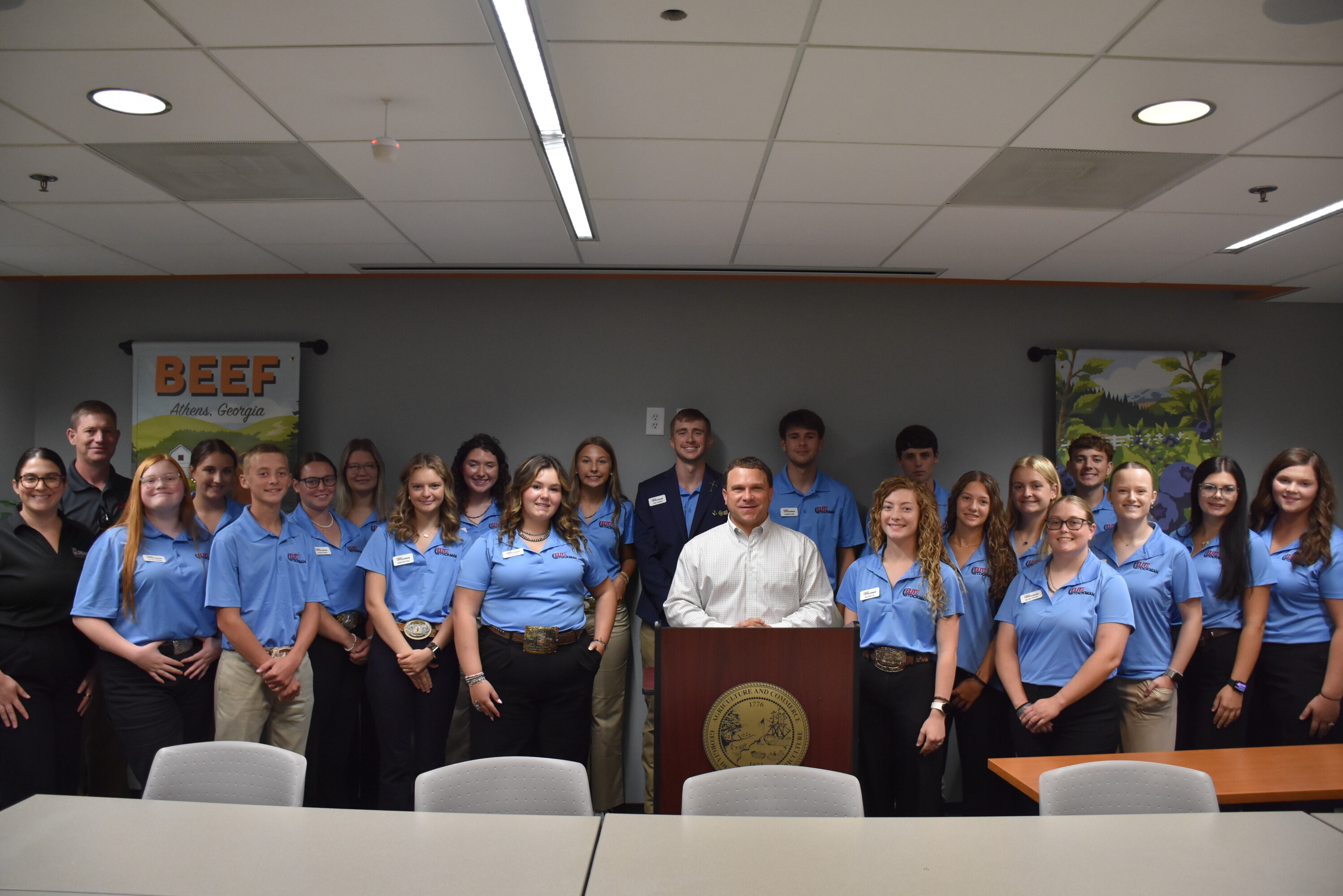 A large group of students in light blue polo shirts pose indoors around Georgia Agriculture Commissioner Tyler Harper, who is standing at a podium bearing the Georgia Department of Agriculture seal